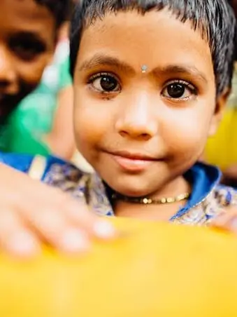 A young child resting on a yellow surface, smiling towards the camera, with other children behind.