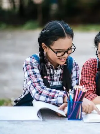 A child in glasses writing in an exercise book outdoors, beside another child.